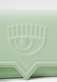 Mintgrön läderyta präglad med en stiliserad ögondesign, med ögonfransar och en cirkulär pupill, ovanför en V-formad sydd kant.