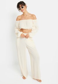 Cremefarbenes, schulterfreies Crop-Top mit langen Ärmeln, kombiniert mit weit geschnittenen Hosen. Der Stoff hat ein leichtes, strukturiertes Aussehen. Accessoires umfassen Sonnenbrillen.