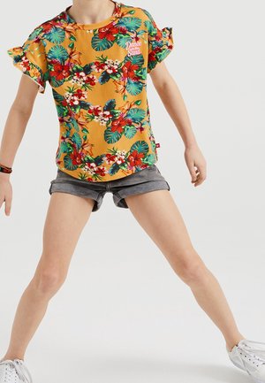 Niño con camiseta amarilla de flores de manga corta y pantalones cortos grises, de pie con las piernas separadas y los brazos ligeramente extendidos.