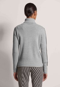 Grauer Strickpullover mit hohem Kragen, geripptem Saum und kontrastierenden Seitenelementen. Trägt dazu gemusterte Hose mit geometrischen Designs.