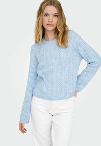 Pull en tricot bleu clair avec un motif texturé, poignets côtelés et col rond, associé à un pantalon blanc.