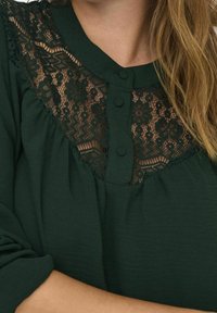 Mörkgrön topp med spetsdetalj och blommönster, rund halsringning och två knappar fram. Mjuk tyg med avslappnad passform.