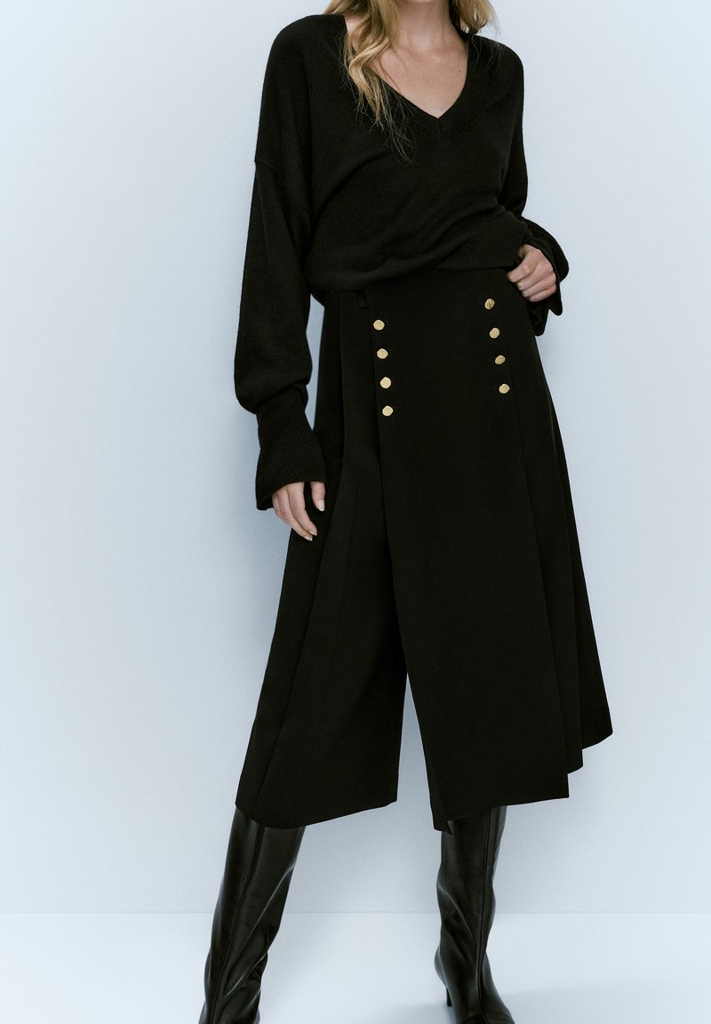 Femme portant un pull noir à col en V, un pantalon noir taille haute à jambes larges avec des boutons dorés, et des bottes noires montant jusqu'aux genoux, debout devant un fond blanc.