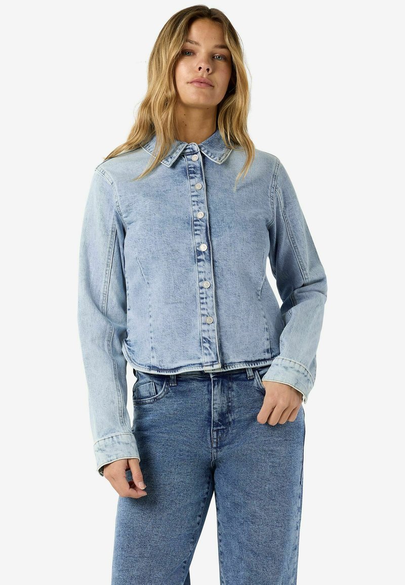 Jeune femme aux longs cheveux blonds portant une veste en jean bleue claire boutonnée et un jean taille haute bleu plus foncé.