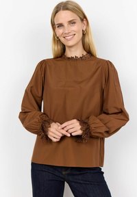 Brown blouse with a high, textured lace collar and puff sleeves. Lace trim at the cuffs adds detail. Paired with dark blue jeans.