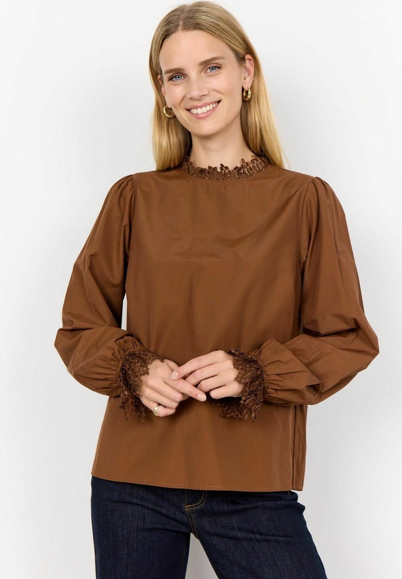 Brown blouse with a high, textured lace collar and puff sleeves. Lace trim at the cuffs adds detail. Paired with dark blue jeans.