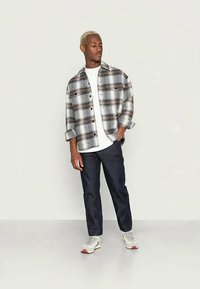 Camisa xadrez em tons de cinza, azul e castanho, com design de botão, mangas enroladas. Combinada com uma t-shirt branca e calças de denim escuras. Ténis brancos.