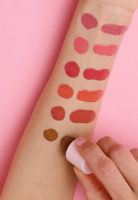 Collezione di rossetti liquidi campionati su un braccio pallido, che mostrano vari toni rossastri e una tonalità marrone, su uno sfondo rosa.