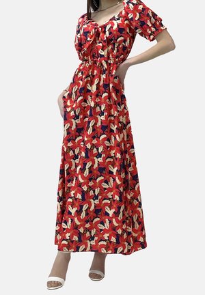 Kobieta ubrana w czerwoną sukienkę maxi w kwiaty z bufkami i wiązaniem z przodu, w parze z białymi sandałami na obcasie z odkrytymi palcami.