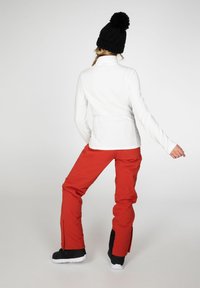 Maglietta bianca a maniche lunghe abbinata a pantaloni rossi brillanti e stivali neri, con cerniere alle caviglie e un berretto nero testurizzato con pom-pom.