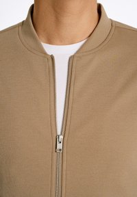 Chaqueta bomber beige con cuello redondo, cremallera frontal y tela texturizada. Superpuesta a una camiseta blanca.