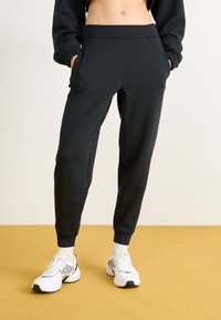 Svarte joggebukser med snøring i livet, sidelommer og smale ben. Laget av mykt stoff med en glatt tekstur og subtile detaljer.