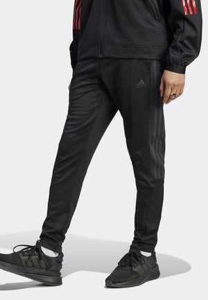 Person trägt schwarze Adidas-Trainingshose mit dezenten drei Streifen und schwarze Sportschuhe vor weißem Hintergrund.
