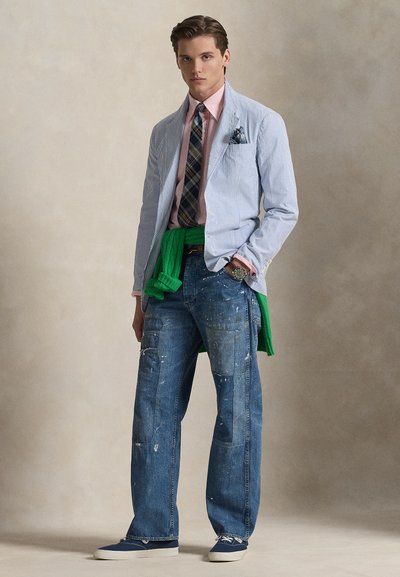 Ριγέ σακάκι με ανοιχτό μπλε και λευκό μοτίβο, ροζ πουκάμισο, γραβάτα καρό, πράσινο πουλόβερ δεμένο στη μέση, σκισμένο μπλε τζιν και αθλητικά παπούτσια.