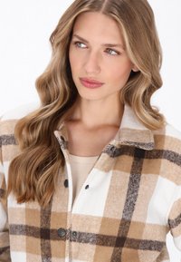 Tan ja valge ruuduline särk-jakk, mis on valmistatud pehmest kangast, kaelusega, mustade nuppudega ja vabalt istuva lõikega.