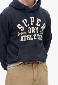 Marineblauwe hoodie met witte tekst "SUPER DRY ATHLETICS Japan Osaka" gedragen met kaki broek en een bruine riem, handen in de capuchonzak.