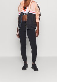 Lichte, kleurgebroken zip-up jack in perzik en zwart, gecombineerd met zwarte tapered broek en zwarte sportschoenen.