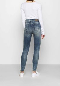 Jeans skinny en denim bleu délavé avec taille haute, dotés de poches arrière et d'une patch en cuir sur la ceinture. Accompagnés de baskets blanches.