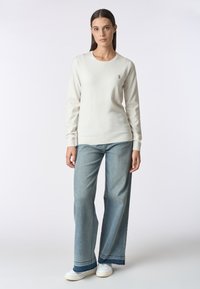 Maglione beige chiaro a maniche lunghe con scollatura rotonda e dettagli ricamati, abbinato a jeans a gamba larga blu e sneakers bianche.