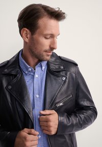 Czarna skórzana kurtka motocyklowa z zatrzaskami i zapinaną na zamek kieszenią, noszona na niebiesko-białej koszuli w kratę z kołnierzykiem i guzikami. Gładka faktura.