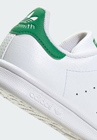 Baltie baltās ādas kedas ar zaļiem akcentiem, kurām ir strukturēta virsma, izšūti sīki sīkumi un zīmols uz papēža un zoles.