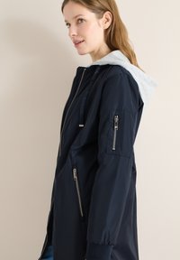 Jeune femme en veste zippée bleu marine avec capuche grise, debout de côté contre un mur beige, regardant légèrement vers le haut et souriant.