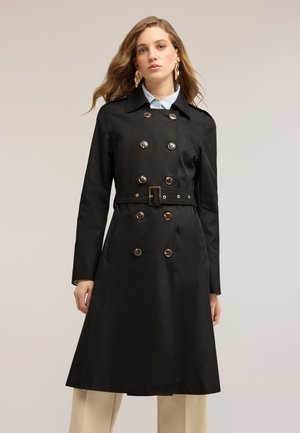 Kvinde iført en sort dobbeltradet trenchcoat med guldknapper og bælte, stående foran en ensfarvet lys baggrund.