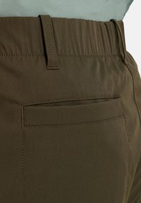 Gros plan sur un pantalon vert olive avec une taille élastique, une passants de ceinture et une poche passepoilée à l'arrière, porté par une personne vêtue d'une chemise gris clair.