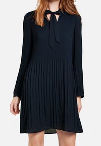 Marinblå plisserad klänning med långa ärmar, v-ringning och knytning vid kragen. Lätt material med en flödande silhuett.