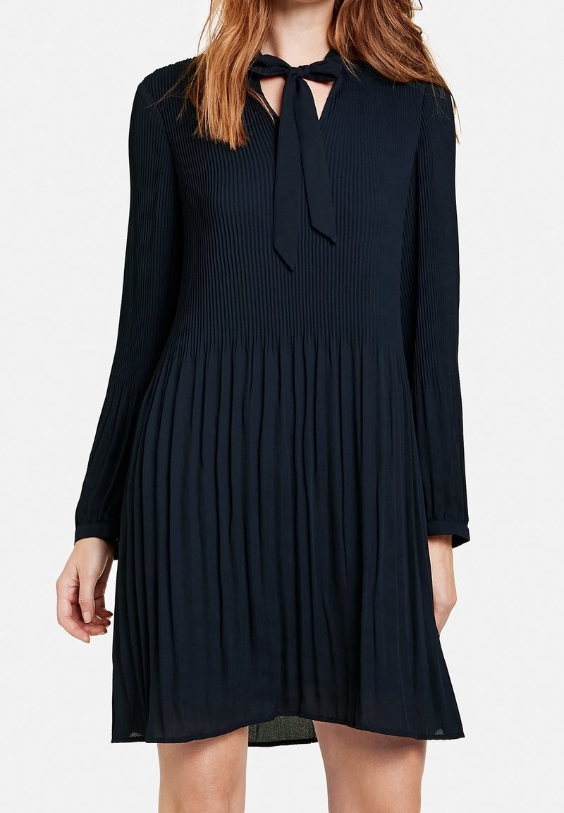 Marinblå plisserad klänning med långa ärmar, v-ringning och knytning vid kragen. Lätt material med en flödande silhuett.