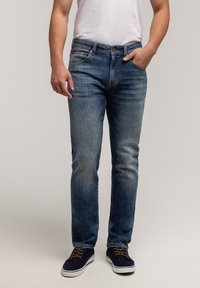 Niebieskie dżinsy o prostym kroju, z wyblakłym wykończeniem, pięcioma kieszeniami i małą czerwoną metką na pasku, zestawione z granatowymi trampkami.