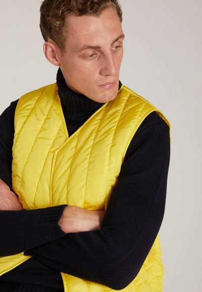 Gelbe Steppweste mit gestepptem Muster, die über einem marineblauen Rollkragenpullover getragen wird. Glatte, glänzende Textur.