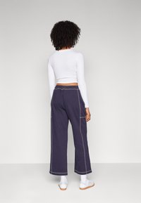 Marineblauwe wijde broek met contrasterende stiksels, gecombineerd met een witte lange mouwen top, gedragen met witte sneakers. Gezichten van achteren.