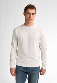 Light cream sweatshirt made of soft fabric, featuring speckles, ribbed cuffs, and a crew neckline. Paired with blue jeans.