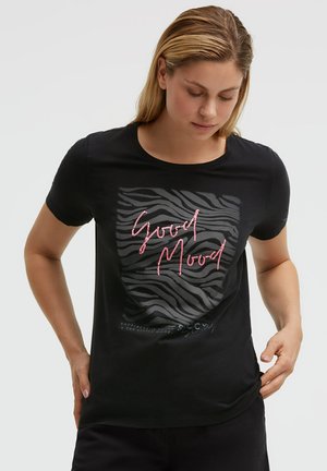 Woman adjusting black T-shirt with dark zebra stripes and pink text reading "Good Mood" on the front against a plain background.