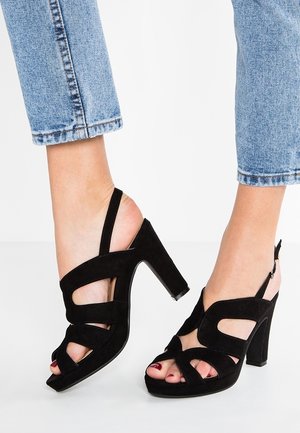 Sandalias de tacón alto de ante negro con puntera abierta, tira trasera ajustable y diseño con recortes. Los tacones presentan una forma gruesa y un acabado texturizado.