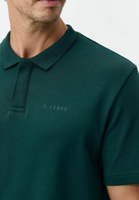 Ciemnozielona koszulka polo wykonana z miękkiej bawełny. Posiada klasyczny kołnierzyk, krótkie rękawy oraz haftowane logo "DIVERSE" w subtelnym odcieniu.