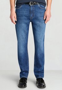 Jeans di denim blu con taglio dritto, caratterizzati dallo stile classico a cinque tasche, leggero scolorimento e cuciture a contrasto, indossati con scarpe nere.