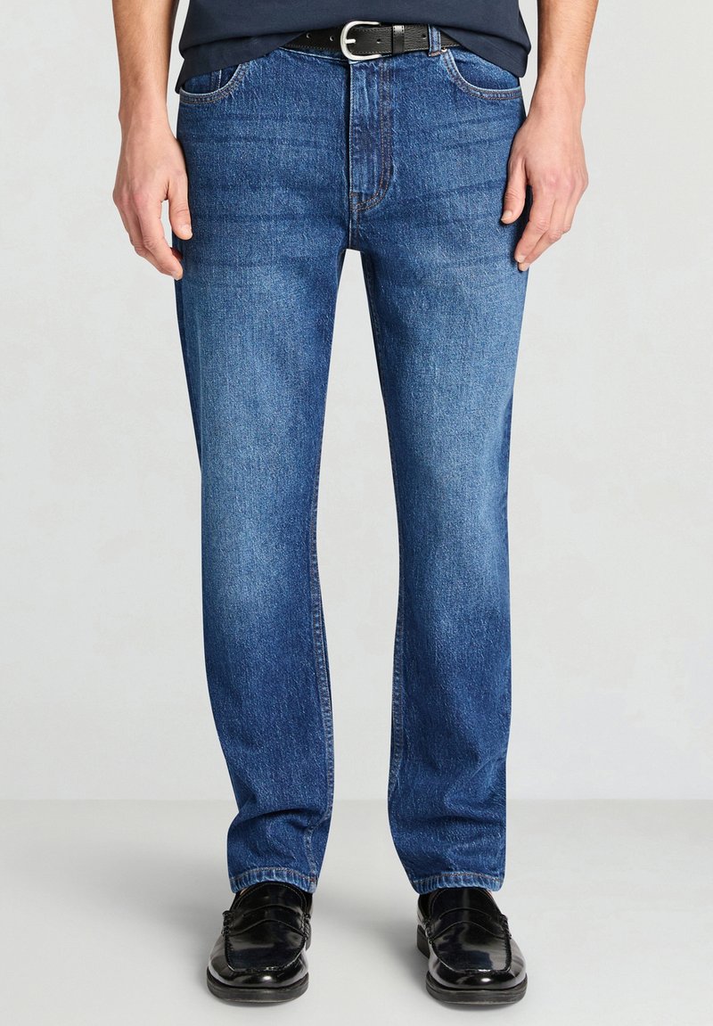 Jeans di denim blu con taglio dritto, caratterizzati dallo stile classico a cinque tasche, leggero scolorimento e cuciture a contrasto, indossati con scarpe nere.