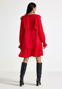 Rød langærmet kjole med flæser ved skuldrene og i kanten, tekstureret stof og parret med sorte knæhøje støvler.