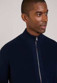 Granatowy sweter z prążkowanej dzianiny z wysokim kołnierzem i srebrnym zamkiem, podkreślający fakturowany wzór i dopasowany krój.