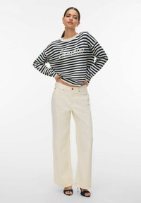 Femme debout, mains sur les hanches, portant un pull à rayures bleu marine et blanc, un pantalon large crème et des talons ouverts noirs sur un fond blanc.