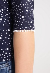 Marineblaue Bluse mit weißen Tupfen, versehen mit einem strukturierten Saum und Pompon-Details. Die Ärmel enden knapp über dem Ellenbogen.