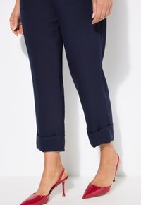 Marinblå tapered-byxor med uppvikta nederkanter, i kombination med glansiga röda slingback-hälar med spetsig tå. Slät textur; inga synliga mönster.
