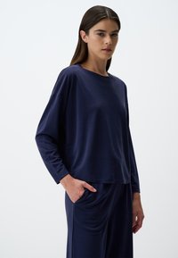 Marineblaues Langarmoberteil aus einem weichen, glatten Stoff. Lässige Passform, Rundhalsausschnitt, mit asymmetrischem Saum und heruntergesetzten Schultern. Kombiniert mit passenden Hosen.