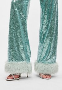 Τουρκουάζ παντελόνια σε φαρδύ κόψιμο με παγιέτες και μπεζ ψεύτικες γούνες στα μανίκια; σε συνδυασμό με ασημένια ψηλοτάκουνα πέδιλα που αναδεικνύουν μια λείη, γυαλιστερή υφή.