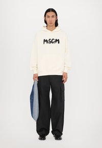 Felpa cremisi con logo "MSGM" nero, abbinata a pantaloni cargo neri larghi. Presenta una borsa di denim tenuta nella mano sinistra. Texture liscia.