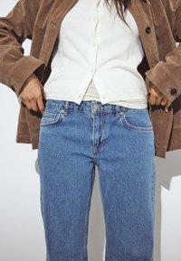 Zilas denima bikses ar taisnu kāju dizainu un smalkām izbalēšanas pazīmēm. Valkātas ar baltu pogu uz augšu kardiganu un brūnu zamšādas jaku.