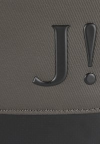 Materiale grigio testurizzato con un logo in rilievo "J!" nero. Presenta dettagli lisci e una cucitura lungo il bordo inferiore.