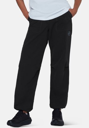 Person iført løstsiddende sorte bukser og sorte sneakers med hænderne i lommerne mod en hvid baggrund.
